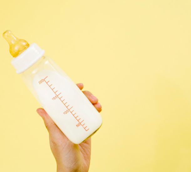 A photo of an adult hand holding a baby bottle filled with milk in front of a yellow background.